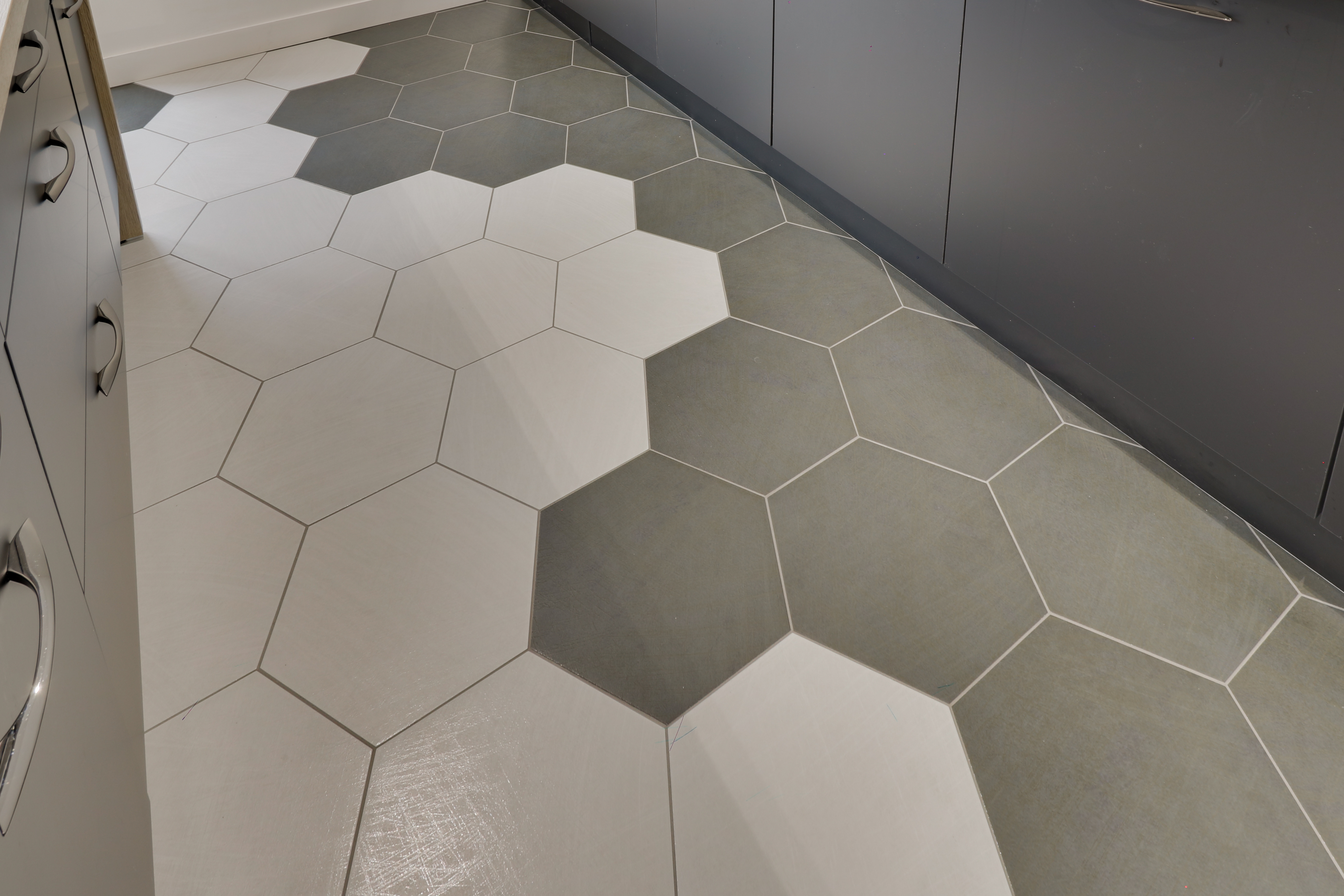 Détail d'un sol de cuisine à carreaux hexagonaux gris et blancs, au pied de meubles gris.